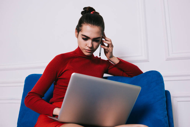 Low angle of young female job seeker speaking on mobile phone and browsing on laptop sitting on blue couch in living room - Photo, Image