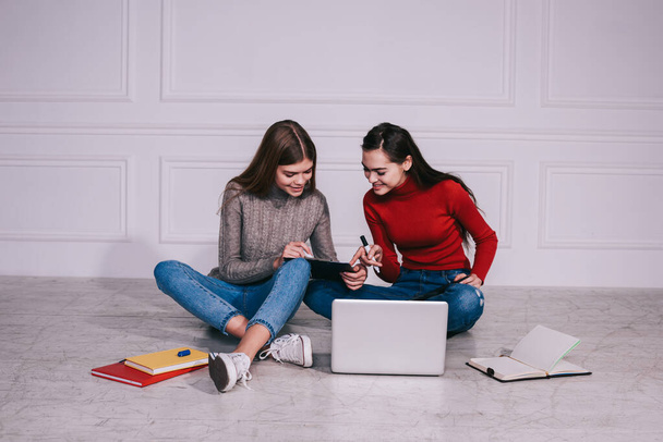 Young female remote workers in casual clothes sitting on floor with laptop and notebooks while working on project together in light room - Photo, image