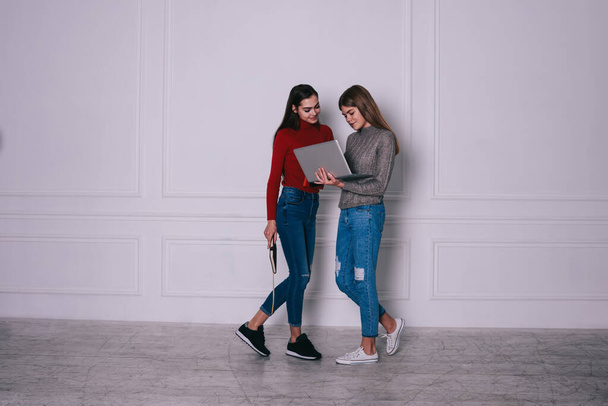 Volledig lichaam van vrolijke jonge vrouwelijke vrienden in casual kleding staan in de buurt van witte muur en browsen laptop in de lichte kamer - Foto, afbeelding