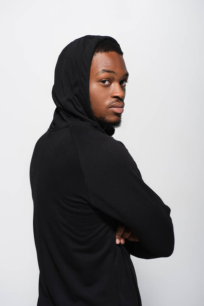 young african american man in hoodie looking at camera while posing with crossed arms isolated on grey - Fotografie, Obrázek