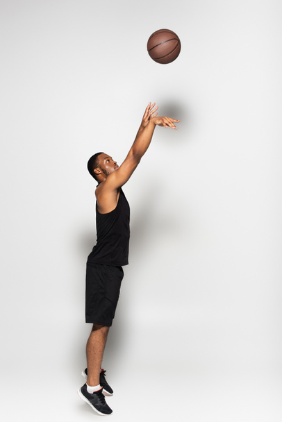 full length of muscular african american man throwing basketball while playing on grey - Фото, зображення