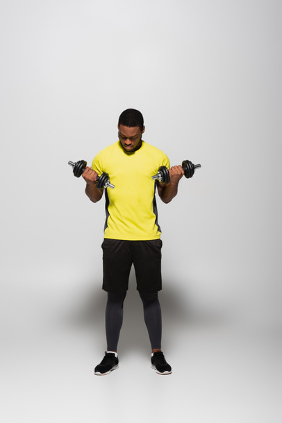 full length of african american sportsman working out with dumbbells on grey  - Photo, Image