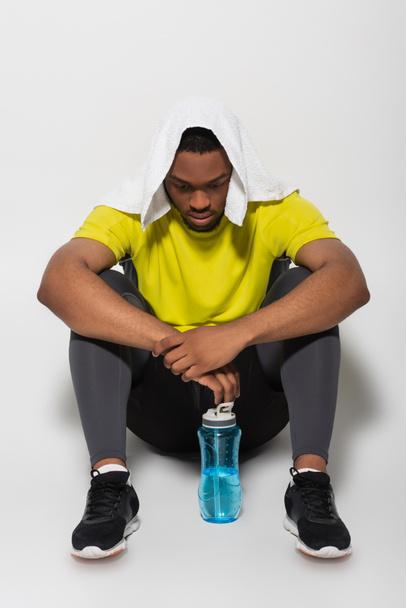 full length of tired african american sportsman with towel on head sitting near sports bottle on grey - Foto, Imagen