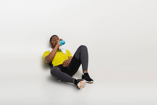 full length of african american sportsman drinking water from sports bottle while sitting on grey - Photo, Image