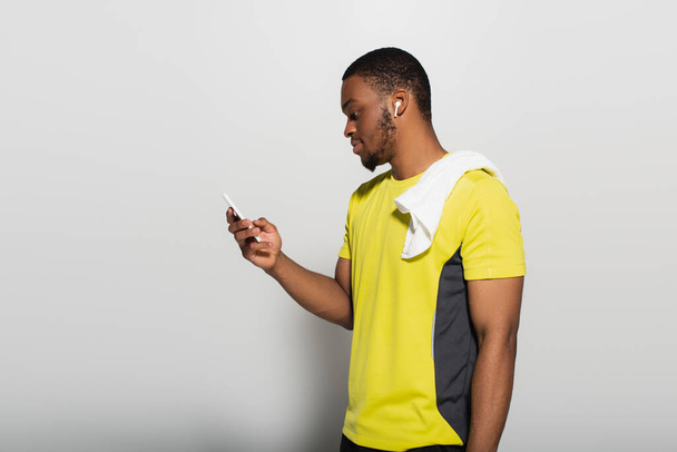 side view of african american sportsman with earphone in ear using smartphone on grey - Foto, Imagem