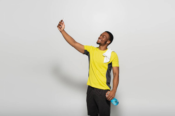 happy african american sportsman with earphone in ear taking selfie on grey - Photo, Image