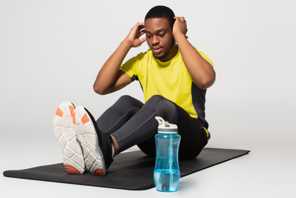 pełna długość sportowy Afroamerykanin człowiek robi mięśnie brzucha ćwiczenia na maty fitness w pobliżu butelki sportowej na szary - Zdjęcie, obraz
