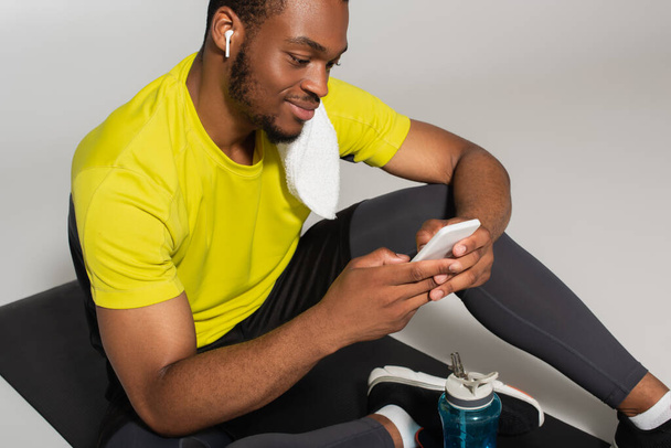 zadowolony afrykański amerykański sportowiec siedzi na macie fitness i za pomocą smartfona w pobliżu butelki sportowej odizolowany na szary  - Zdjęcie, obraz