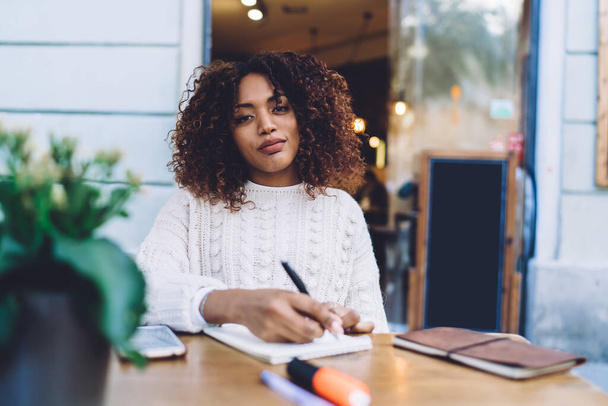 Positive African American female in casual clothes sitting at wooden table with smartphone and taking notes in copybook while resting in cafe - Foto, Imagem