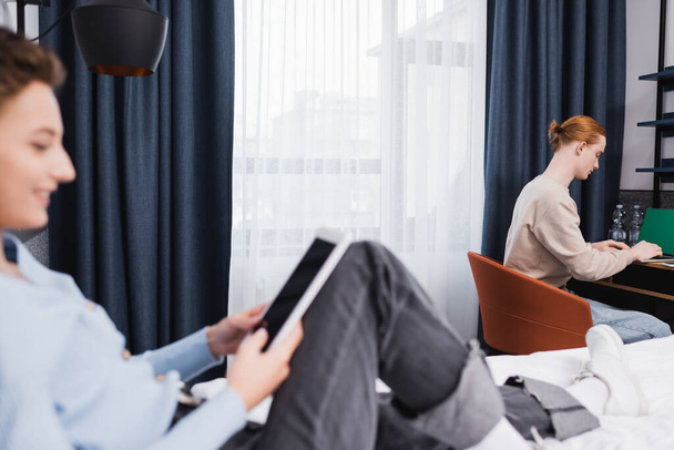 Вид сбоку на человека, использующего ноутбук рядом с размытой подругой с цифровой табличкой в гостиничном номере  - Фото, изображение