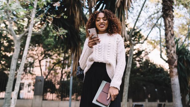 Beautiful dark skinned female in trendy wear reading messages and notification on smartphone spending time in town, positive african american woman blogger satisfied with successful content - Foto, Imagem