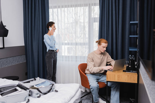Smiling woman talking on smartphone near boyfriend using laptop and suitcase on bed in hotel  - Foto, Bild