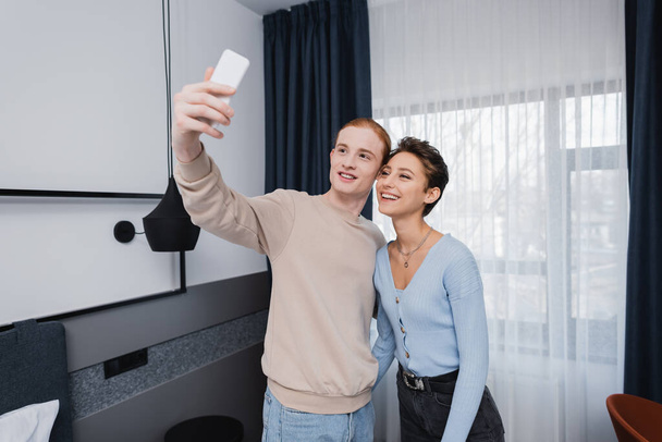 Glimlachend stel neemt selfie op smartphone in moderne hotelkamer  - Foto, afbeelding