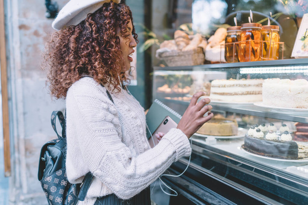 Side view of positive African American female in casual clothes and beret with backpack standing on street and choosing dessert from window shop while listening to music - Zdjęcie, obraz