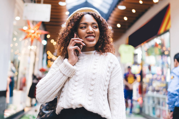 Happy African American female in casual clothes and beret standing in modern mall and talking on smartphone during shopping and looking away - 写真・画像