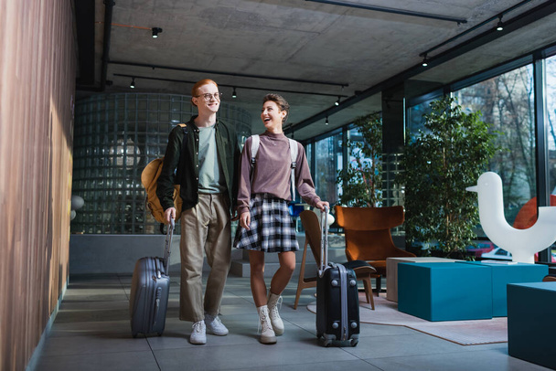 Cheerful woman with backpack and suitcase walking near boyfriend in hotel lobby  - Photo, Image