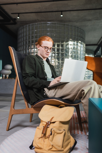 Redhead man using laptop near suitcase in hotel lobby  - Фото, зображення