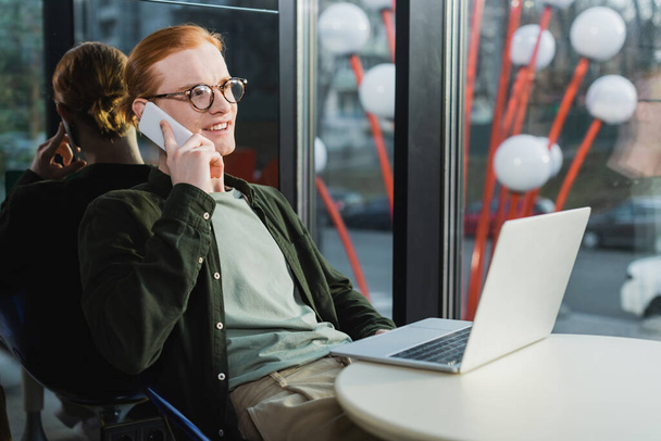 Ευτυχισμένος κοκκινομάλλης ελεύθερος επαγγελματίας μιλώντας στο smartphone κοντά στο laptop στο λόμπι του ξενοδοχείου  - Φωτογραφία, εικόνα