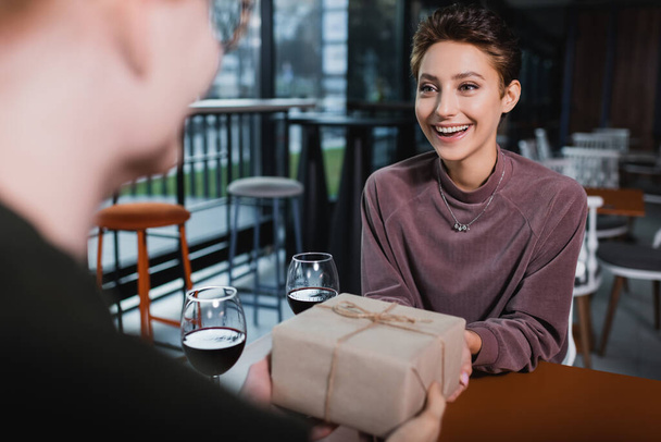Excited woman holding present near wine and blurred boyfriend in hotel cafe  - Photo, Image