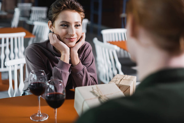 Νεαρή γυναίκα κοιτάζοντας θολή φίλο με δώρο κοντά στο κρασί στο καφέ του ξενοδοχείου  - Φωτογραφία, εικόνα