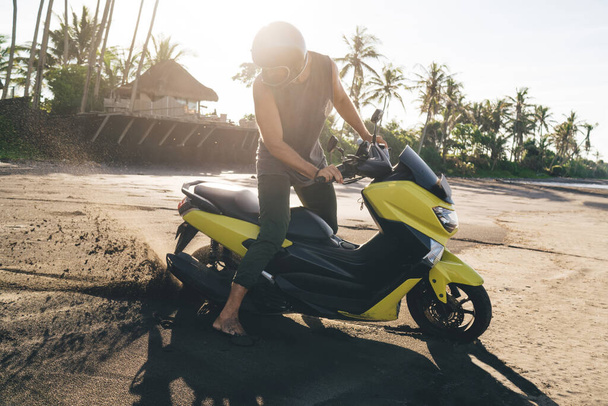 Side view of young anonymous male traveler in casual clothes and helmet sitting on modern motorbike on sandy beach in tropical resort - Photo, image
