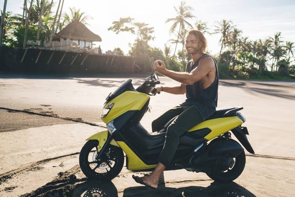 Full body of happy male hipster in undershirt sitting on motorcycle and chilling in tropical country while enjoying summer alone - Foto, afbeelding