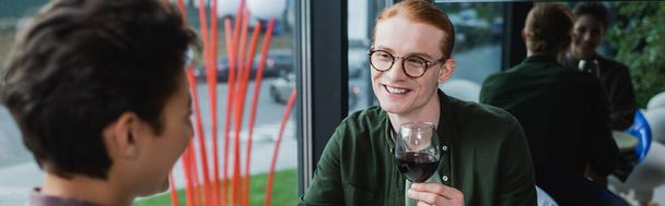 Smiling red haired man holding glass of wine near blurred woman in hotel, banner  - Photo, Image