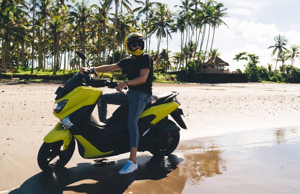 Stylish male biker in casual wear and helmet sitting on motorbike and admiring beauty of ocean against scenic palm trees landscape with blue sky on sunny summer day - Фото, изображение