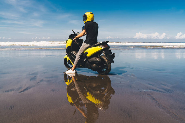 Side view of unrecognizable stylish guy in helmet and casual outfit sitting on trendy motorcycle parked on sandy beach near powerful ocean against blue sky - Photo, Image