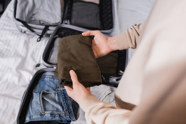 Cropped view of man holding clothes near blurred suitcases on hotel bed  - Foto, imagen