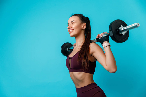 beautiful sportive brunette woman holding a barbell on her shoulders on a blue background. isolated - Photo, Image