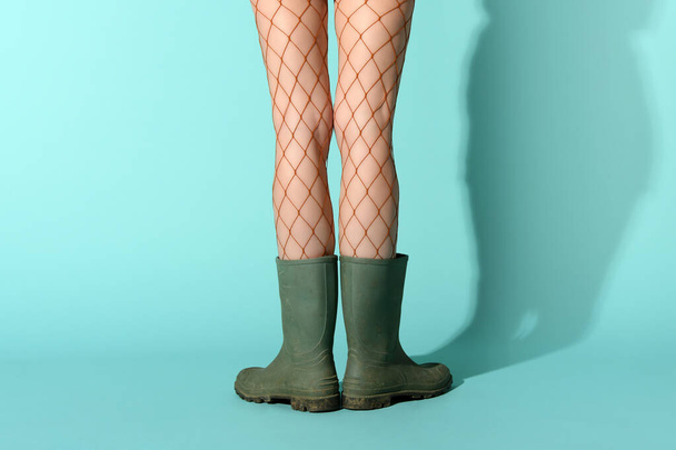 Crop unrecognizable female model wearing fishnet stockings and dirty rubber boots standing in blue studio - 写真・画像