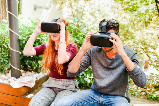 Young couple sitting on wooden bench and experiencing another virtual world in headset in park against green plants - Foto, immagini