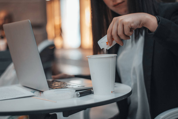 Joven gerente de trabajo vertiendo azúcar en el café en una cafetería en la plaza de la ciudad mientras trabaja con su computadora portátil - Foto, Imagen