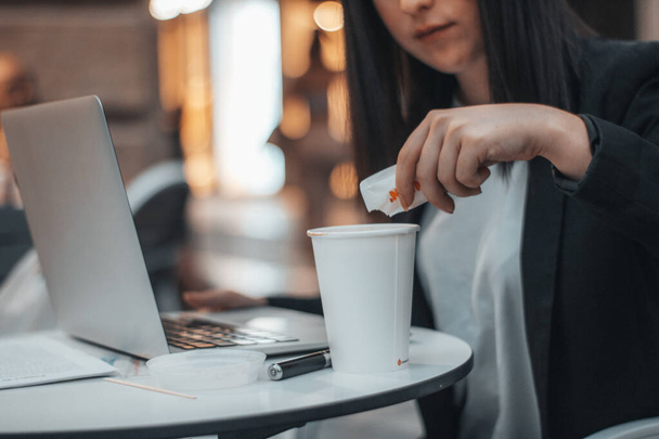 Joven gerente de trabajo vertiendo azúcar en el café en una cafetería en la plaza de la ciudad mientras trabaja con su computadora portátil - Foto, imagen
