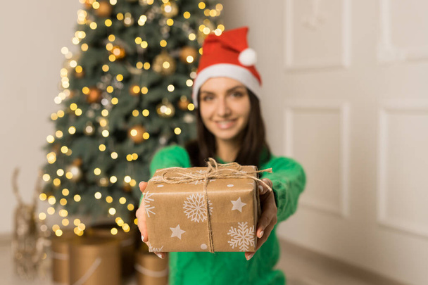 Das junge lächelnde Mädchen hält ein braunes Geschenk in der Nähe des Weihnachtsbaums - Foto, Bild