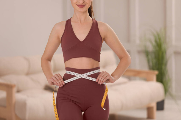 Happy woman in sportswear measuring waist with tape at home, closeup - Fotografie, Obrázek