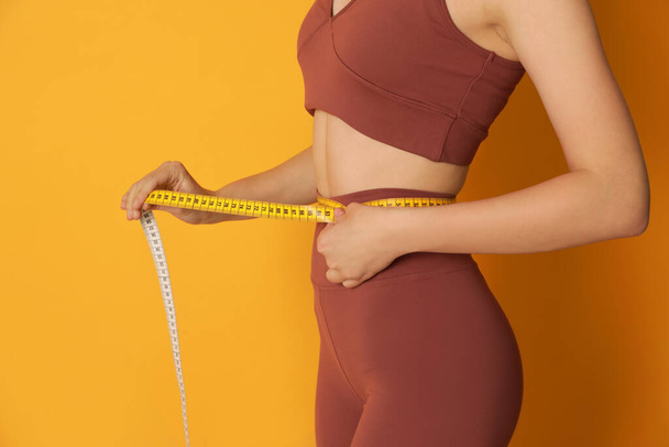Woman in sportswear measuring waist with tape on yellow background, closeup - Фото, изображение