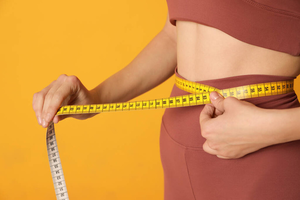 Woman in sportswear measuring waist with tape on yellow background, closeup - Zdjęcie, obraz