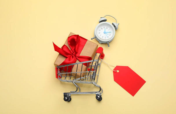 Shopping cart with tag, gift box and alarm clock on beige background, flat lay - Valokuva, kuva