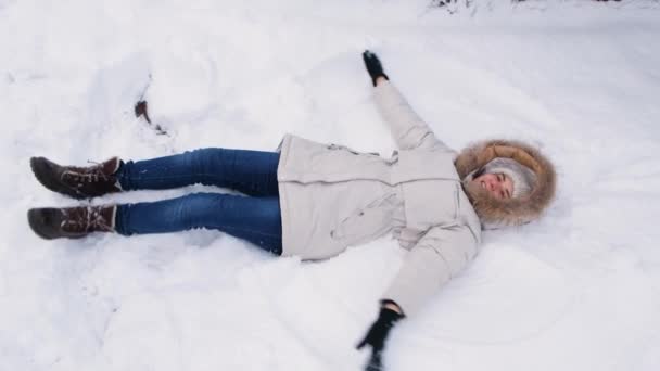 Beyaz ceketli bir kadın karda uzanıyor, ellerini kaldırıyor, sanki kanatlarını çırpıyor ve bir melek yapıyor. - Video, Çekim