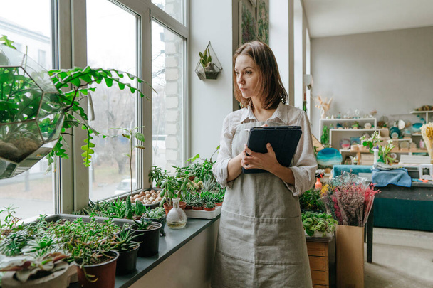 Eine junge Floristin mit Schürze steht in ihrer Pflanzenwerkstatt und hält Tabletten in der Hand. Selektiver Fokus. - Foto, Bild