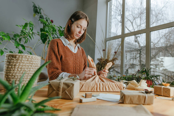 Young woman packing gift box in fabric furoshiki style at her workshop. Eco friendly package concept. Selective focus. - Foto, Bild