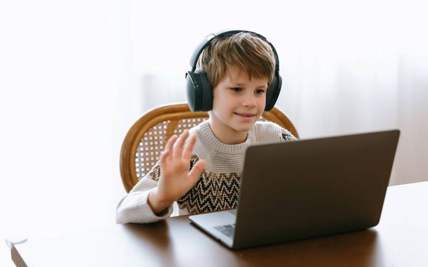 Porträt eines niedlichen kleinen Jungen mit Headset und winkt am Bildschirm während einer Videokonferenz am Laptop mit dem Lehrer von zu Hause im Wintertag. Neue Normalität, soziale Distanzierung und Daheimbleiben. - Foto, Bild