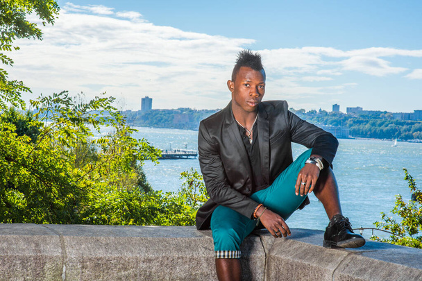 Man Relaxing Outside. Dressing in a black blazer,  green pants, a young black guy with mohawk hair is sitting on a rocky stone fence by a river, relaxing. Urban Fashion. - Photo, image