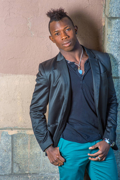 Dressing in a black blazer, green pants, a young black guy with mohawk hair is standing against the wall, his head tilted one side, confidently looking at you - Photo, Image
