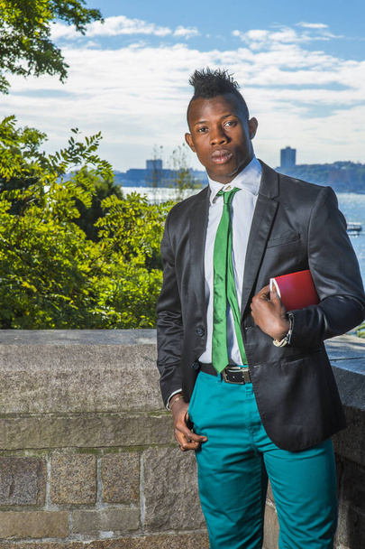 Modern Businessman. Wearing a black blazer, green pants, green neck tie, a hand holding a red book under arm, a young black guy with mohawk haircut is standing by a river, looking at you - Photo, Image
