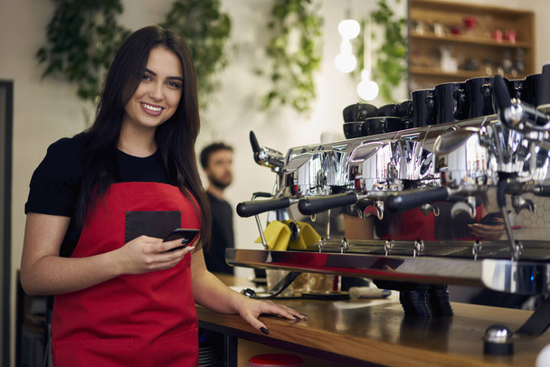 Professional brunette woman in uniform standing at bar in cafeteria with mobile phone chatting in blog with followers, half length millennial portrait of female barista enjoying job at coffee shop - Foto, immagini