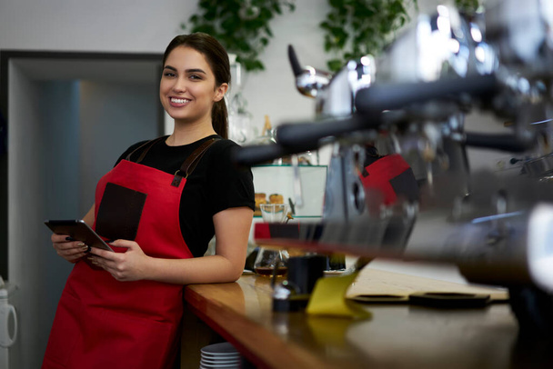 Half length portrait of cheerful woman barista in red apron enjoying working process using modern digital table for checking accountings, skilled female waitress at working place in near equipment - Fotoğraf, Görsel
