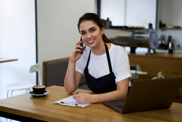 Smiling caucasian woman waitress talking on mobile phone about service delivery making notes in form, skilled professional female manager making smartphone call to banking service checking income - Photo, Image